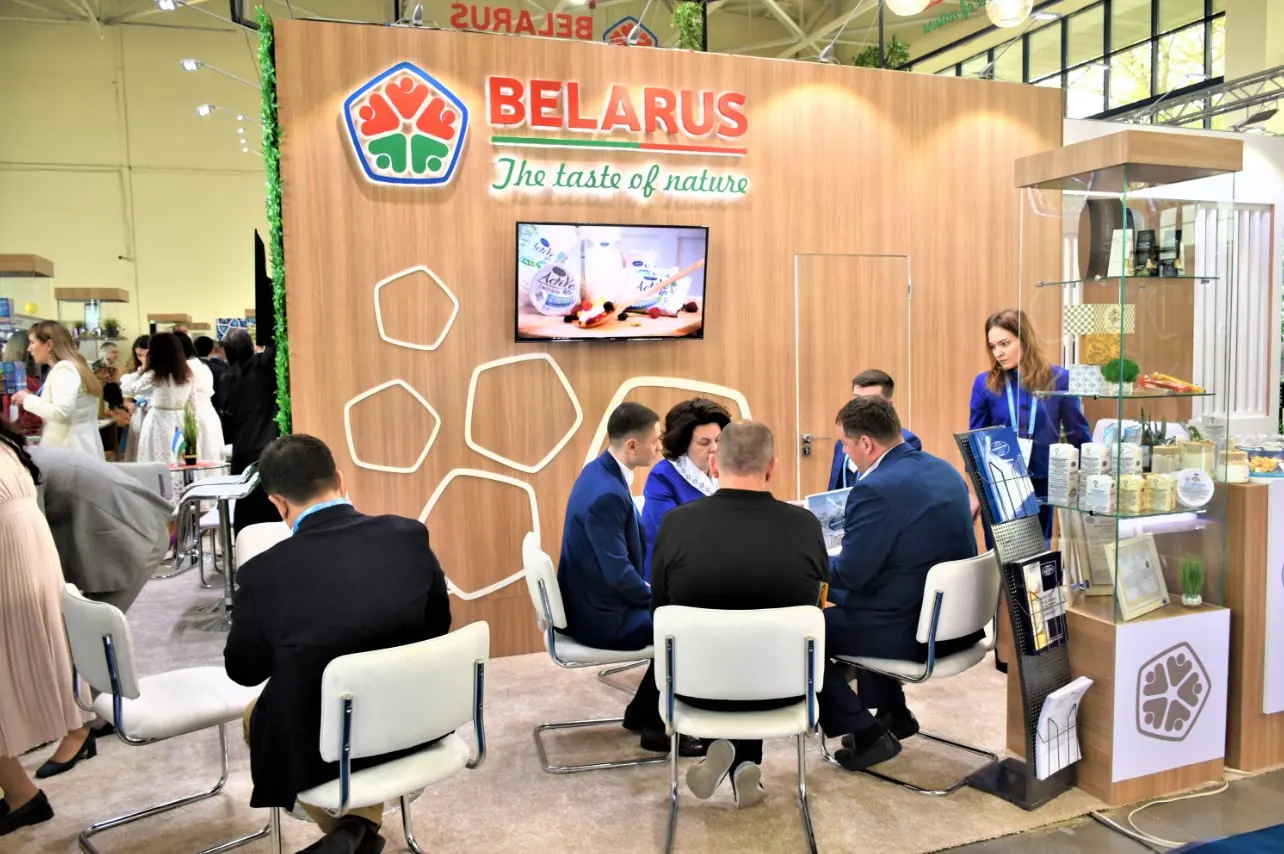 Выставочный стенд Belarus с деревянной отделкой, крупным логотипом, белыми шестигранниками, экраном и переговорной зоной.